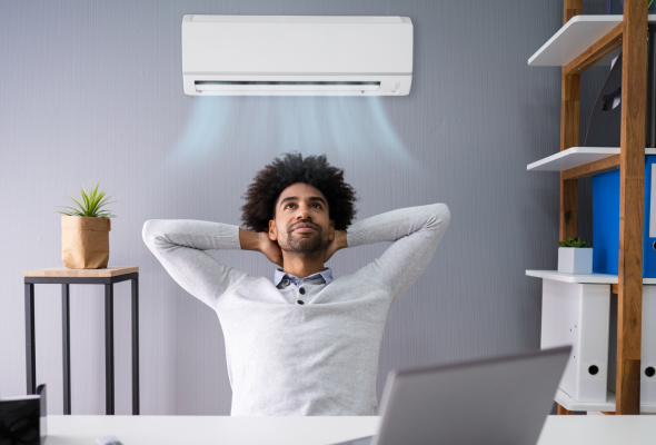 A man sat at a desk with his arms up behind his head in a relaxing position. An air conditioning unit behind him blowing out cold air waves. he looks comfortable with his laptop on his desk.