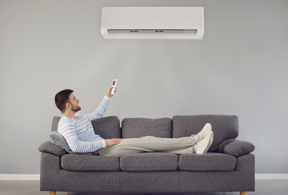 A man sat on her sofa with his feet up. He is pointing a remote in the direction of a wall mounted air conditioning unit.