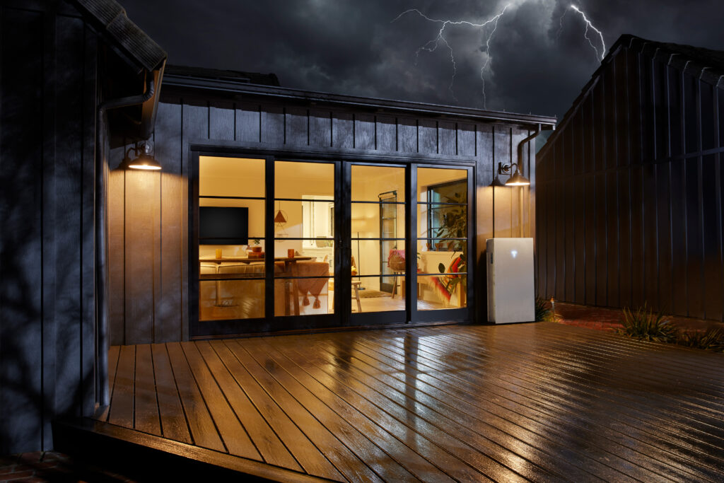 An image of a house with bi folds, the the lights on inside and a Tesla Powerwall 3 on the outside wall. A Dark grey cloudy sky above the house.