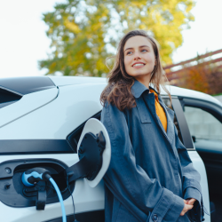 A lady stood next to her car which is plugged into a charging port. she is smiling.