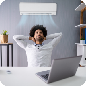 A man sat at a desk with his hands behind his head in a comfortable position. A wall mounted air conditioning unit behind him blowing out air.