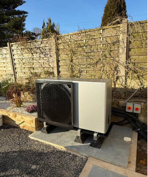 image of an Air Source Heat Pump in a garden with fence panels behind and blue skies above.