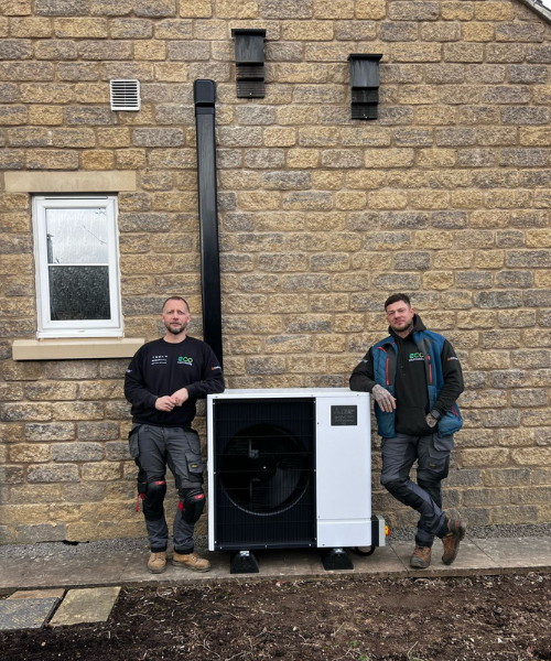 Image of an Air Source Heat Pump up against a stone wall with 1 man stood at one side and one man stood at the other side. Both men have Eco Partners uniform on.