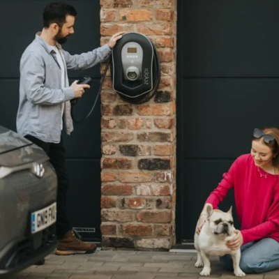 Image of a man and woman. The man is stood holding a Zappi EV Charging cable. And the woman is kneeling down with a small dog. Both next to a car and a garage door.